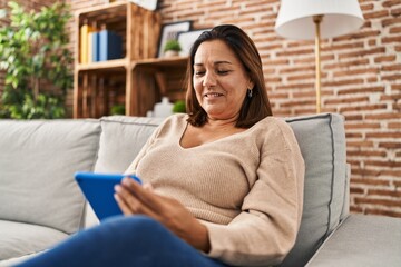 Middle age hispanic woman using touchpad sitting on sofa at home