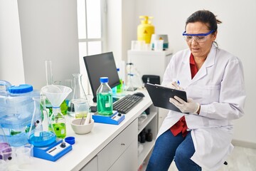 Middle age hispanic woman wearing scientist uniform writing on clipboard at laboratory