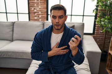 Young hispanic man using inhaler sitting on sofa at home