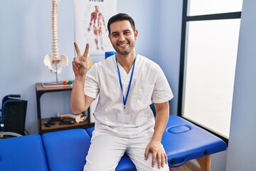 Young hispanic man with beard working at pain recovery clinic showing and pointing up with fingers number two while smiling confident and happy.