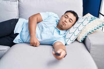 Young chinese man watching tv lying on sofa with boring expression at home