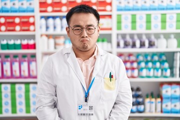 Chinese young man working at pharmacy drugstore puffing cheeks with funny face. mouth inflated with air, crazy expression.