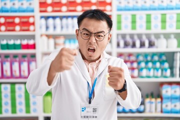 Chinese young man working at pharmacy drugstore angry and mad raising fists frustrated and furious while shouting with anger. rage and aggressive concept.