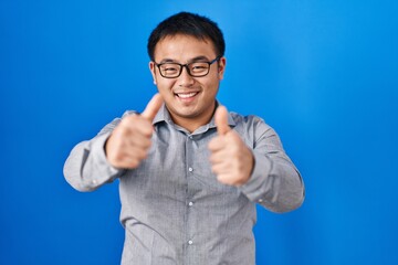 Naklejka premium Young chinese man standing over blue background approving doing positive gesture with hand, thumbs up smiling and happy for success. winner gesture.