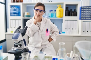 Fototapeta premium Hispanic girl with down syndrome working at scientist laboratory with hand on chin thinking about question, pensive expression. smiling with thoughtful face. doubt concept.