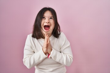 Woman with down syndrome standing over pink background begging and praying with hands together with hope expression on face very emotional and worried. begging.