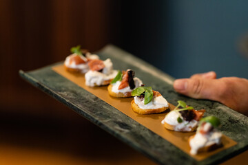 horizontal shot of server's hand holding dark serving tray of crostini with ricotta prosciutto and basil
