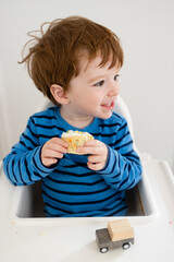 Cute caucasian toddler boy in blue and black striped shirt sits at white highchair eating a vanilla cupcake with sprinkles on his birthday