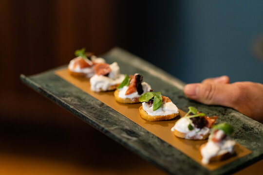 horizontal shot of server's hand holding dark serving tray of crostini with ricotta prosciutto and basil
