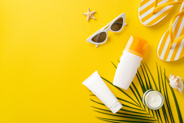 Sunbathing supplies concept. Top view arrangement showcasing sunscreen bottles without labels, sunglasses, flip-flops, shell, starfish, palm leaf on vivid yellow backdrop with space for text or promo