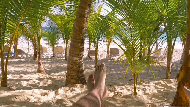 Man lying in a sunbed in palm tree's shadow on a beach. Close up young man feet resting on beach on a summer holiday. POV feet of young man lying on a sandy beach by the ocean. Summer holiday concept.
