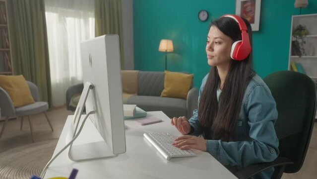 A woman in headphones works at a computer. A young woman listens to music, and types on the keyboard. Profile portrait of an Asian woman at a table with a computer. HDR BT2020 HLG Material.
