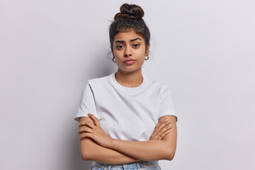 Studio shot of serious Indian woman with dark hair combed in bun keeps arms folded waits for explanations listens attentively information dressed in basic t shirt isolated over white background. - 614558500