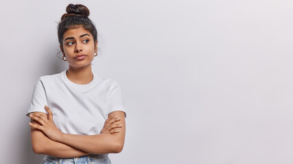 Studio shot of pleasant looking Iranian woman keeps arms folded concentrated aside with thoughtful expression wears casual t shirt isolated over white background copy space for your advertisement - 614559577