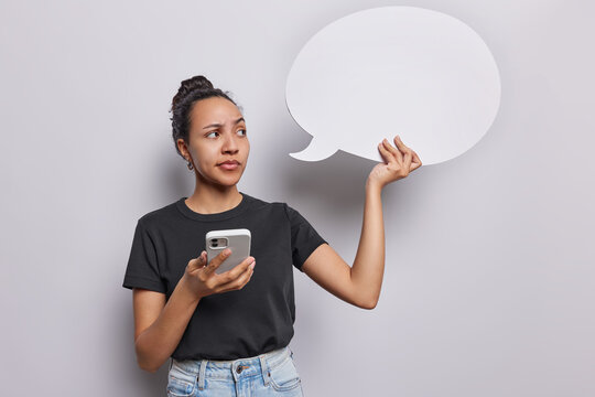 Horizontal shot of pretty thoughtful Latin woman holds speech bubble and modern cellular concentrated aside wears casual black t shirt and jeans isolated on white background. Place your promotion here