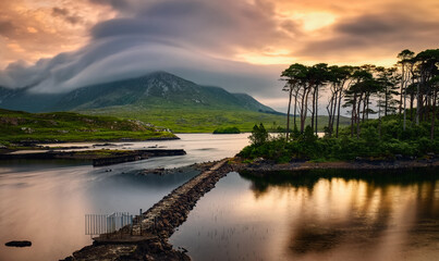 Obraz premium Dramatic cloudy sunrise lakeside landscape scenery of twelve pines island reflected in water surrounded by mountains at Derryclare, connemara national Park in County Galway, Ireland 