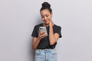 Pleased young Latin woman glances at her smartphone engrossed in checking sms has focused gaze and faint glow of screen smiles gently dressed in casual black t shirt and jeans keeps hand on neck