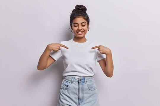 Young dark haired woman captures attention with her vibrant energy with contagious smile directs focus to her blank white tshirt using two fingers shows place for your logo or design poses indoor