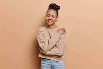 Calm dark haired woman embracing herself with self assuredness her smile reflecting genuine pleasure and self love keeps eyes closed dressed in casual pullover and jeans isolated over brown background