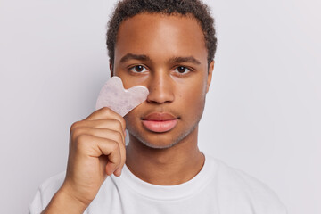 Headshot of dark skinned man uses gua sha stone for massaging wears casual t shirt concentrated...