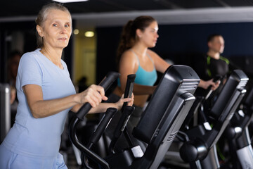 Fototapeta na wymiar Concentrated elderly woman working out on elliptical machine in gym. Concept of healthy lifestyle of older generation