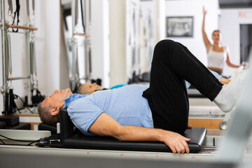 Mature male pensioner in sportswear performing exercise lying down on reformer bed during back training in Pilates center