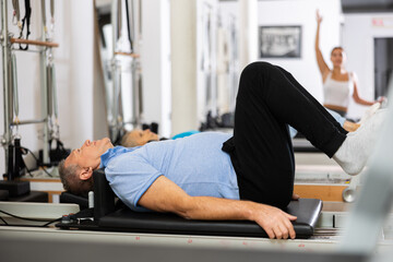 Fototapeta na wymiar Mature male pensioner in sportswear performing exercise lying down on reformer bed during back training in Pilates center