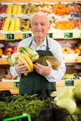 Caucasian old man in apron standing in greengrocer with fruits and vegetables in hands and looking in camera.