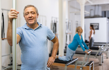 Fototapeta na wymiar Portrait of smiling elderly man in activewear posing against women exercising on reformer bed machine in Pilates studio