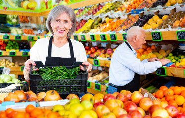 Senior woman and man working in salesroom of greengrocer. Woman holding crate full of green pepper, man setting out goods on shelves behind her.
