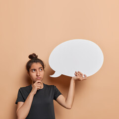 Discontent thoughtful Latin woman holds chin concentrated above being deep in thoughts holds blank communication bubble for your promotional text wears black t shirt isolated over brown background