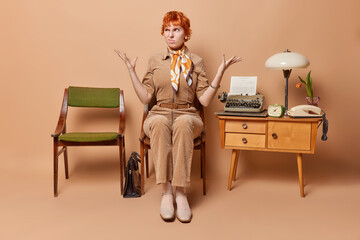 Irritated redhead young woman raises hands and expresses annoyance poses on chair near table with...