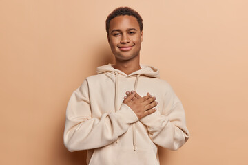 Dark skinned handsome adult man with palm pressed against his chest symbolizes essence of love expresses gratitude for received compliment wears casual sweatshirt isolated over brown background