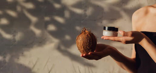 Close-up of woman hands holding coconut cream for skin at textured wall backdrop. Arms of young lady with cosmetic cream in shadow sunlight. Concept of cosmetology and self care. Copy ad text space - Powered by Adobe