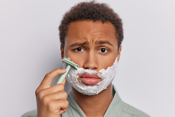 Headshot of dark skinned African man with curly short hair applies shaving foam on his beard wears bathrobe isolated over white background. Male beauty routine concept. Morning daily hygiene