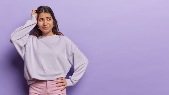 Horizontal shot of confused Latin woman scratches head focused aside tries to make decision wears casual pullover and pink trousers isolated over purple background copy space for your advertisement