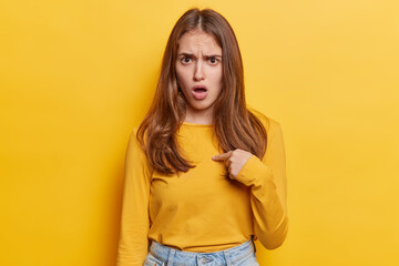 Young woman points finger at chest verbally defending herself with perplexed and puzzled expression...
