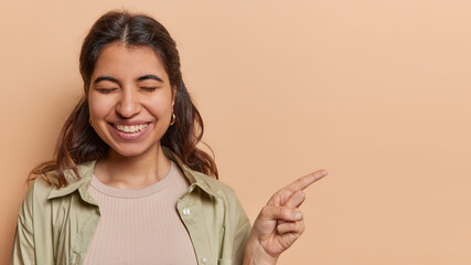 Horizontal shot of dark haired young Latin woman smiles pleasantly points index finger on copy space shows advertising promo deal keeps eyes closed wears shirt isolated over brown background