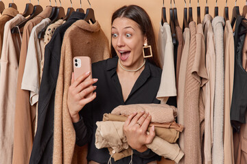 Shocked brunette woman stares in smartphone holds folded clothes finds out about big discounts poses next to selection of clothes on hangers eagerly seeks new outfit to purchase reveals amazing offer