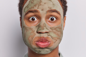 Close up image of stunned young man meticulously applies beauty clay mask to his face has breath caught in amazement blows cheeks stands speechless isolated over white background. Beauty concept