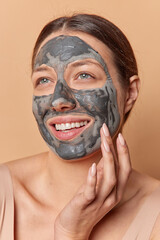 Vertical shot of dark haired woman applies nourishing clay mask for skin care takes care of overall wellbeing smiles pleasantly wears t shirt isolated over brown background. Rejuvenation procedures