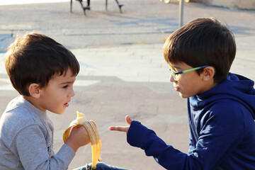 Two brothers sitting on a bench talking while one eats a banana.