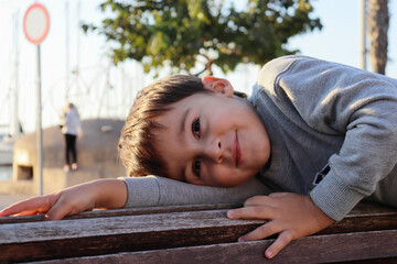Five year old boy in gray pullover resting and smiling lying on a wooden bench. Horizontal image.