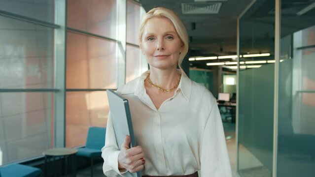 Serious business woman going with clipboard in business center hallway. Portrait of businesswoman CEO in office corridor looking at camera 4K. Close up mature confident woman 50s walking in corridor