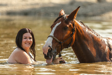 A young woman swimming with her bay brown horse in a pond in summer outdoors