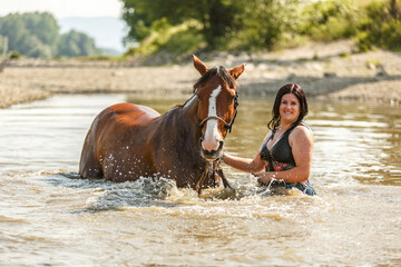 A young woman swimming with her bay brown horse in a pond in summer outdoors