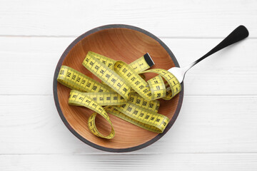 Bowl with measuring tape and fork on white wooden table, flat lay. Weight loss concept