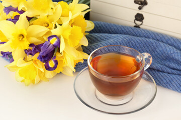 Cup of aromatic tea, beautiful yellow daffodil and iris flowers on white table