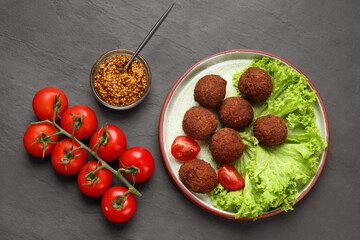 Delicious falafel balls, tomatoes, lettuce and grain mustard on dark grey textured table, flat lay. Vegan meat products
