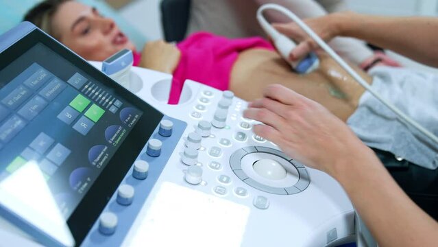Ultrasound machine is used to check up the pregnancy development. Doctor moves the device by the belly of blonde woman.